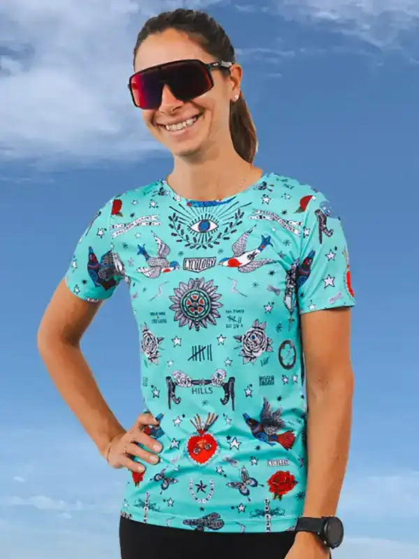Woman wearing vibrant turquoise cycling jersey with tattoo-style graphics, sunglasses, and watch against blue sky