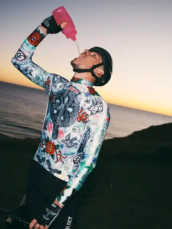 Cyclist drinking from pink bottle in tattoo-print cycling jersey at sunset, ocean view