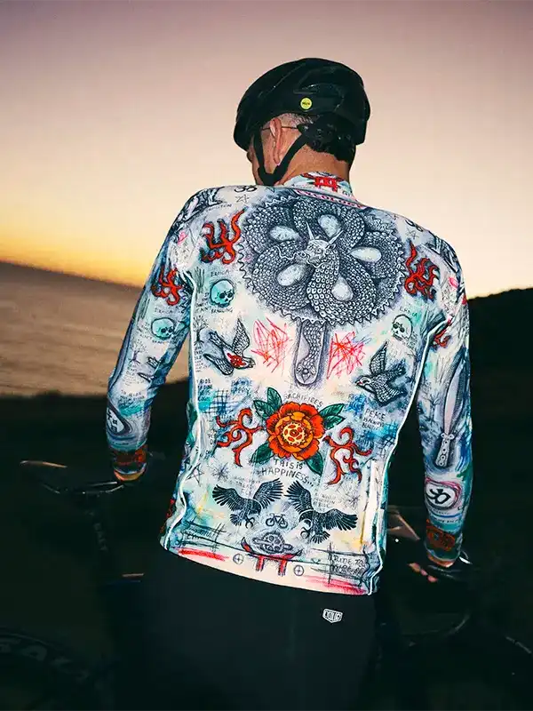 Men's cycling jersey with tattoo-style graphics, worn at sunset near ocean, featuring skulls, roses, and birds