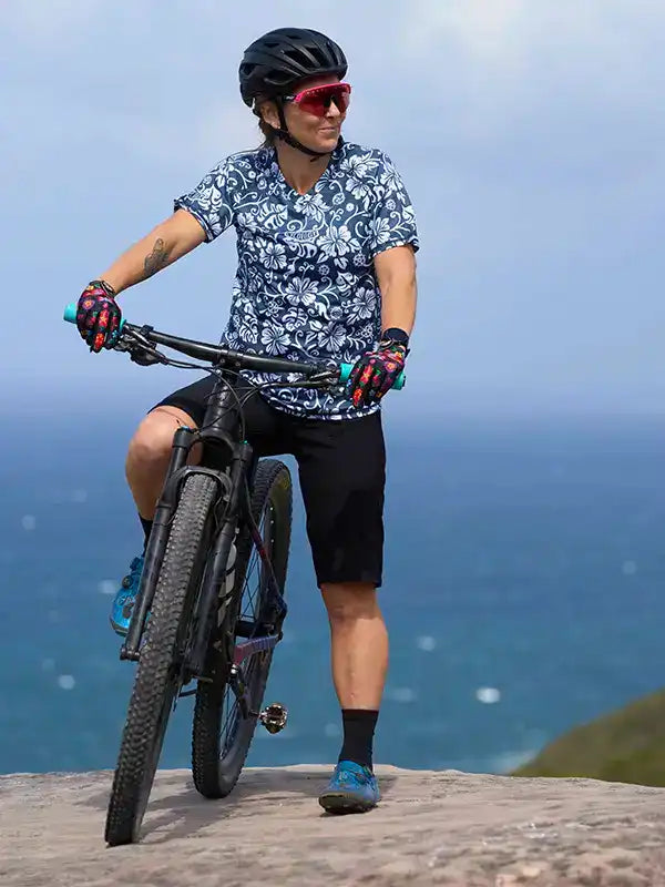 Cyclist in floral cycling jersey and black shorts riding mountain bike by ocean, wearing helmet and gloves