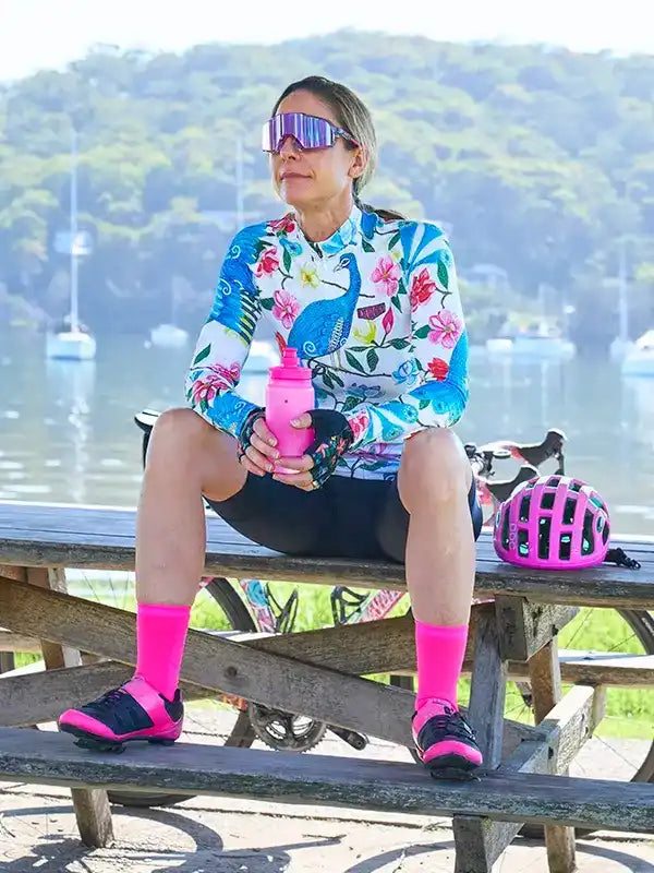 Woman in floral cycling jersey and pink socks sitting by water with bike helmet and water bottle