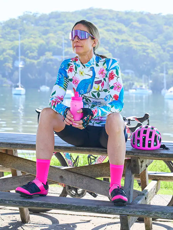 Woman sitting on a picnic table with a scenic background, wearing a White Women's Long Sleeve Summer Cycling Jersey with peacock and floral pattern on a white background