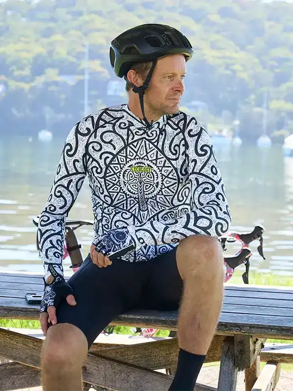 Man wearing patterned cycling jersey and helmet, sitting on bench by water, ready for ride