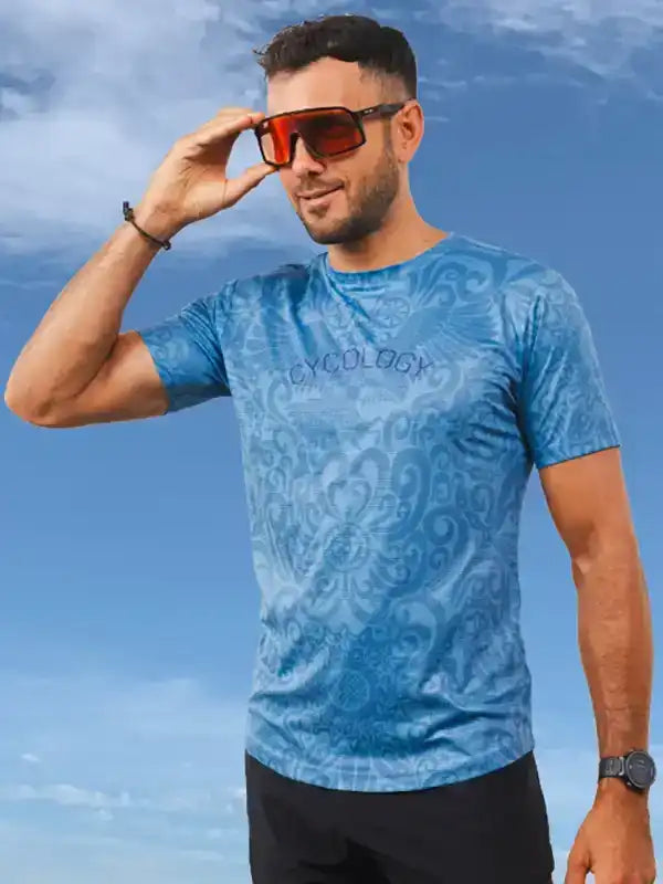 Man wearing blue patterned cycling jersey with 'CYCOLOGY' text, sunglasses, against sky background