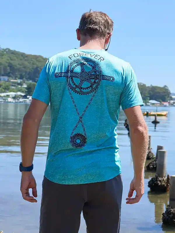 Man wearing turquoise cycling jersey with 'FOREVER' and gear graphic, standing by water