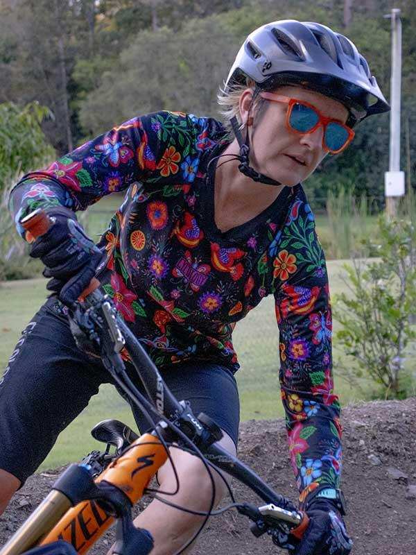 Woman cycling in vibrant floral long-sleeve jersey, helmet, and sunglasses on dirt trail