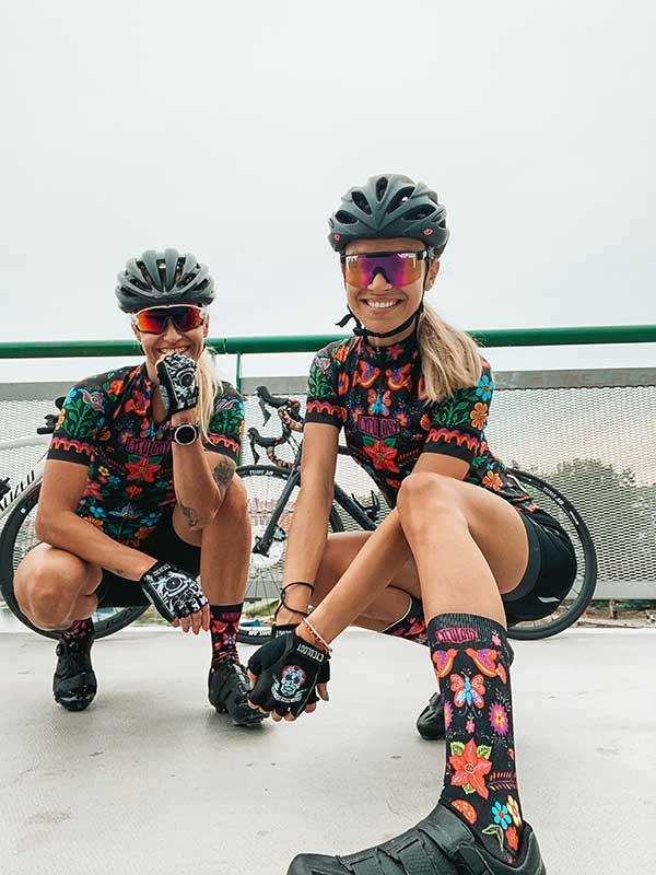 Women in colorful floral cycling jerseys, helmets, and socks posing with bikes outdoors