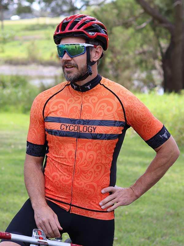 Man wearing orange patterned cycling jersey with CYCOLOGY logo, helmet, and sunglasses outdoors