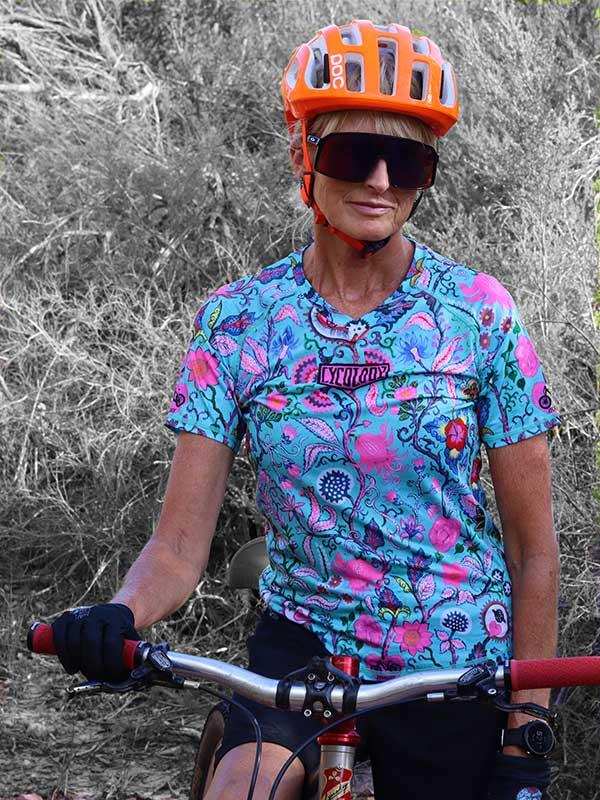 Woman cycling in vibrant floral jersey, orange helmet, and sunglasses on trail