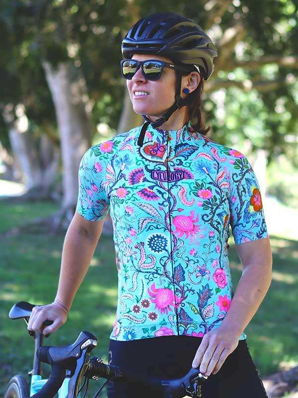 Woman in vibrant floral cycling jersey, helmet, and sunglasses, holding bike outdoors