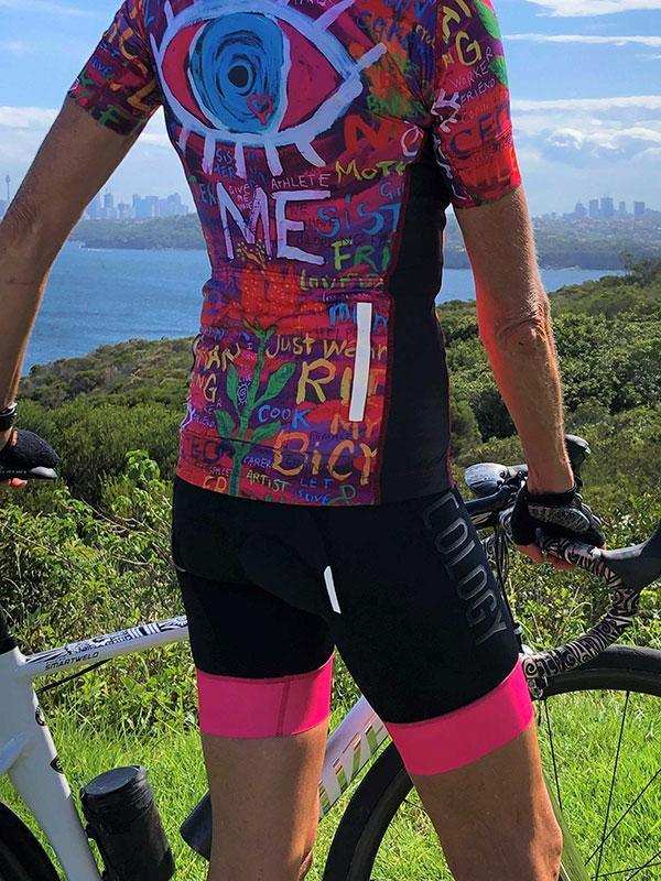Colorful graffiti-style cycling jersey with bold graphics, worn with black shorts and pink leg bands, outdoors near water.
