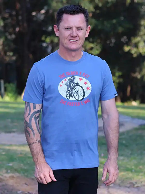 Man wearing blue cycling t-shirt with cyclist graphic and 'The Older I Get The Faster I Was' text outdoors