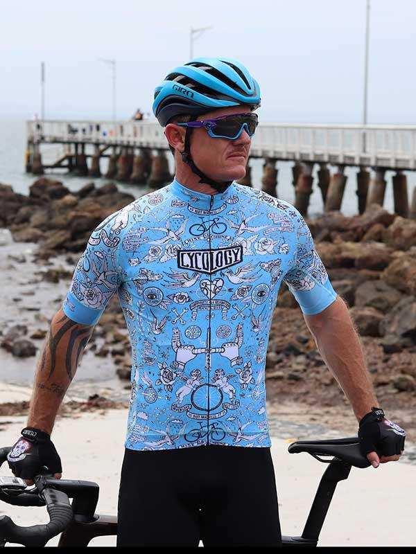 Man in blue patterned cycling jersey and helmet by seaside pier, holding bike
